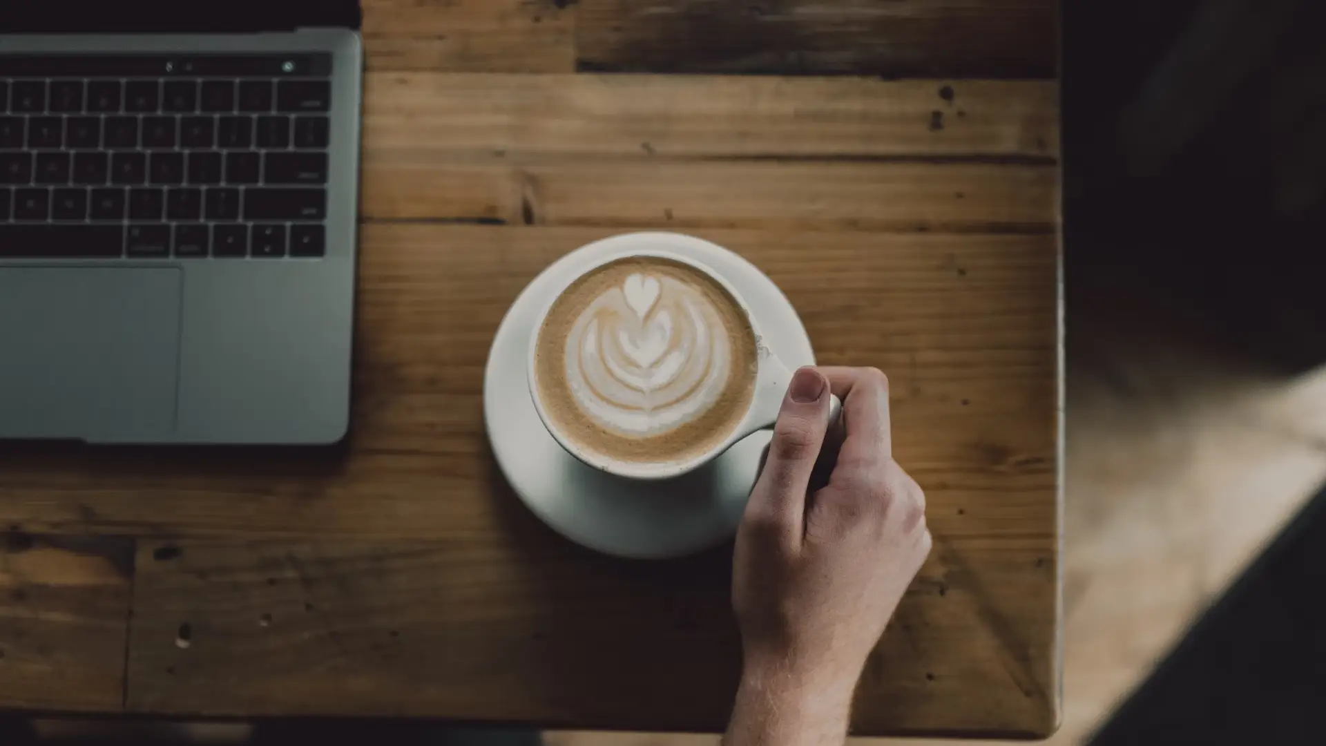 A Cup with Latte Art on a Brown Desk with a Laptop in the Corner