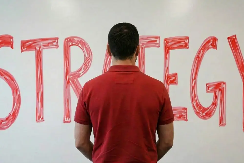Man Standing in front of Strategy White Board A man in a red polo is standing in front of a white board that says "Strategy"