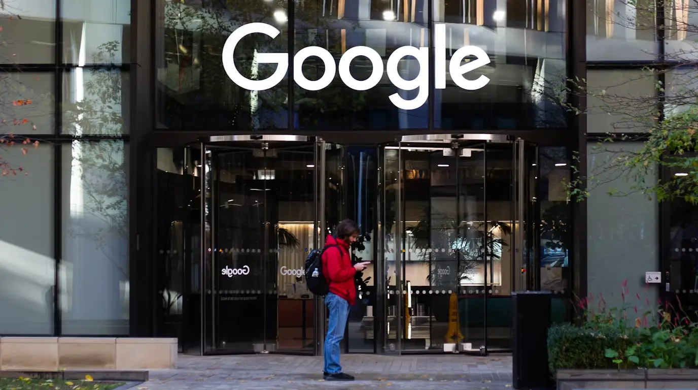 Google sign on a new glass building with a person in a red jacket and blue jeans standing outside.