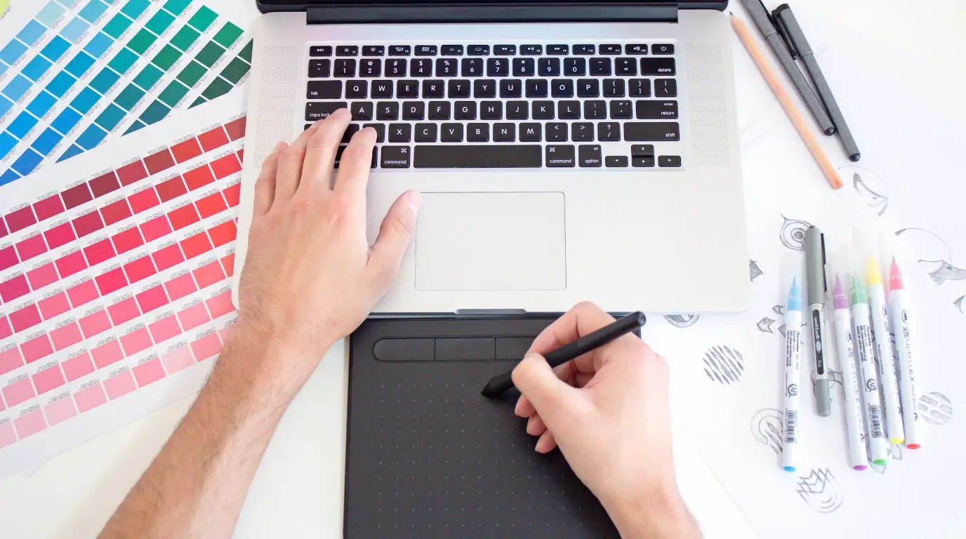 A MacBook keyboard and a drawing pad on a desk with color swatches to the right and a hand reaching forward.