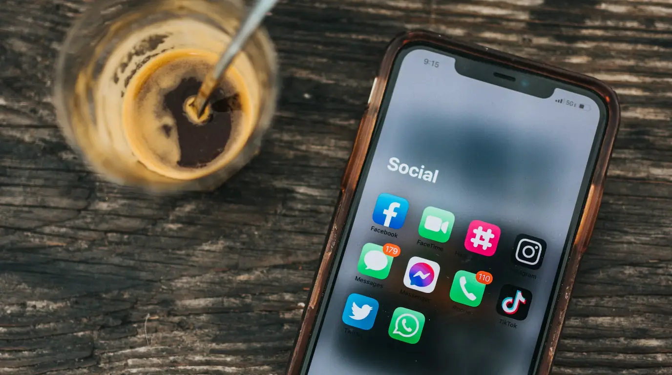 Social media icons in an iPhone folder that is sitting on a rustic wooden desk with coffee in the corner.