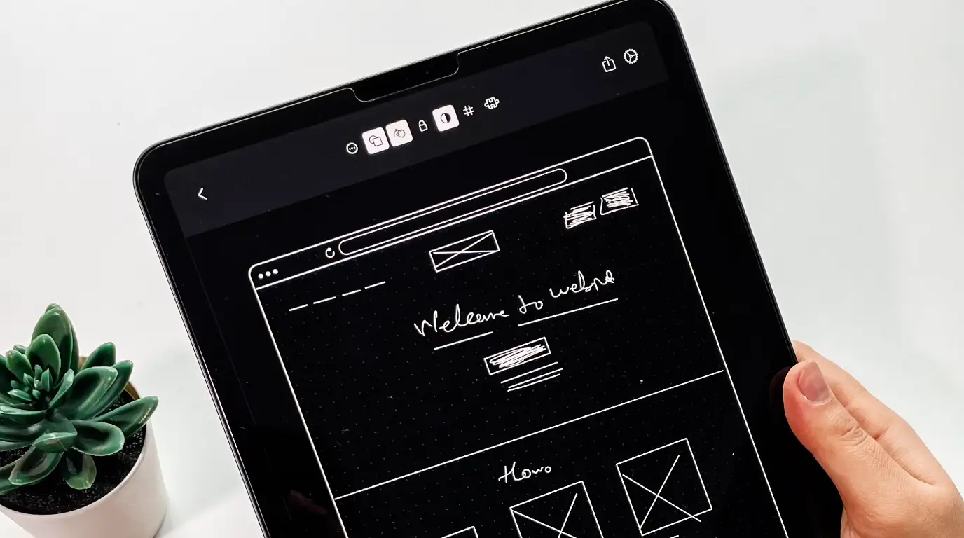 A sketch of a website wireframe on an iPad with a small plant on a white desk in the backrgound.
